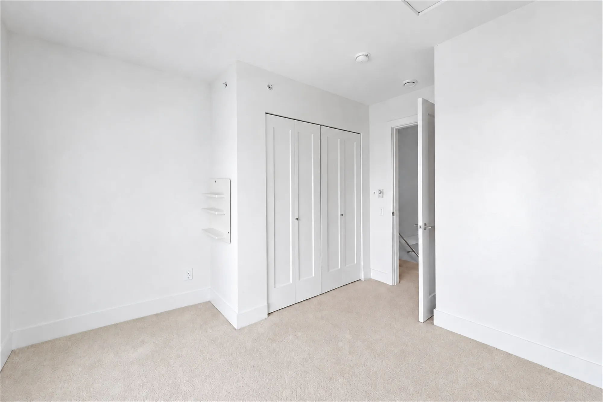 The back bedroom shown empty: the double closet with bifold doors, the door to the hall, a small wall-mounted shelf, carpet