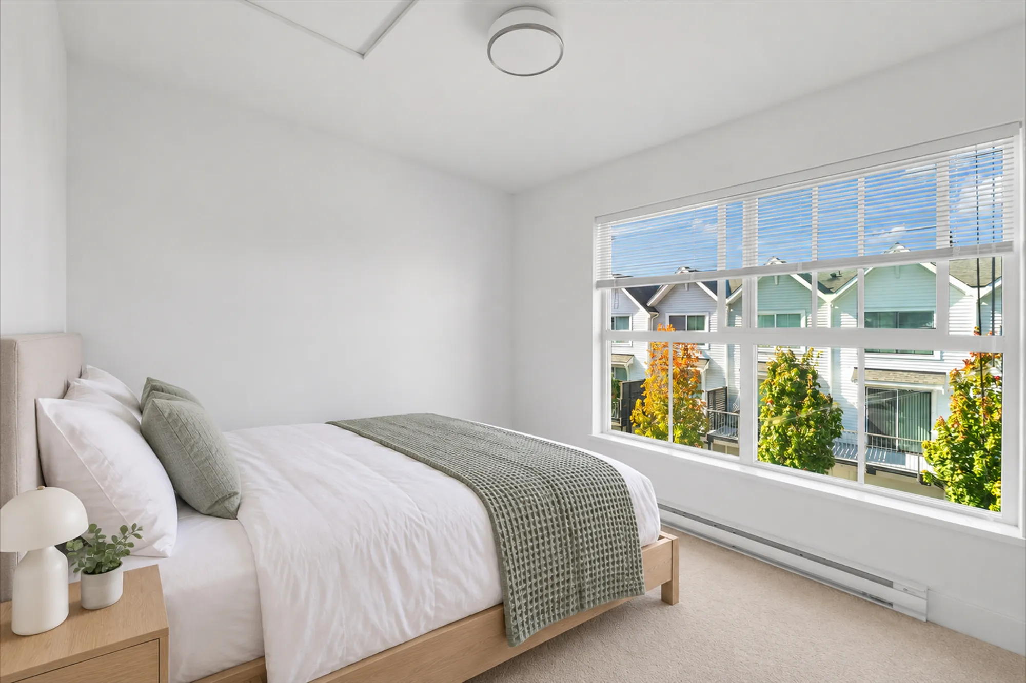 The back bedroom virtually staged: a wide window with blinds over the lane, a bed with white linen and a green throw, a night table with a lamp and a plant, a baseboard heater