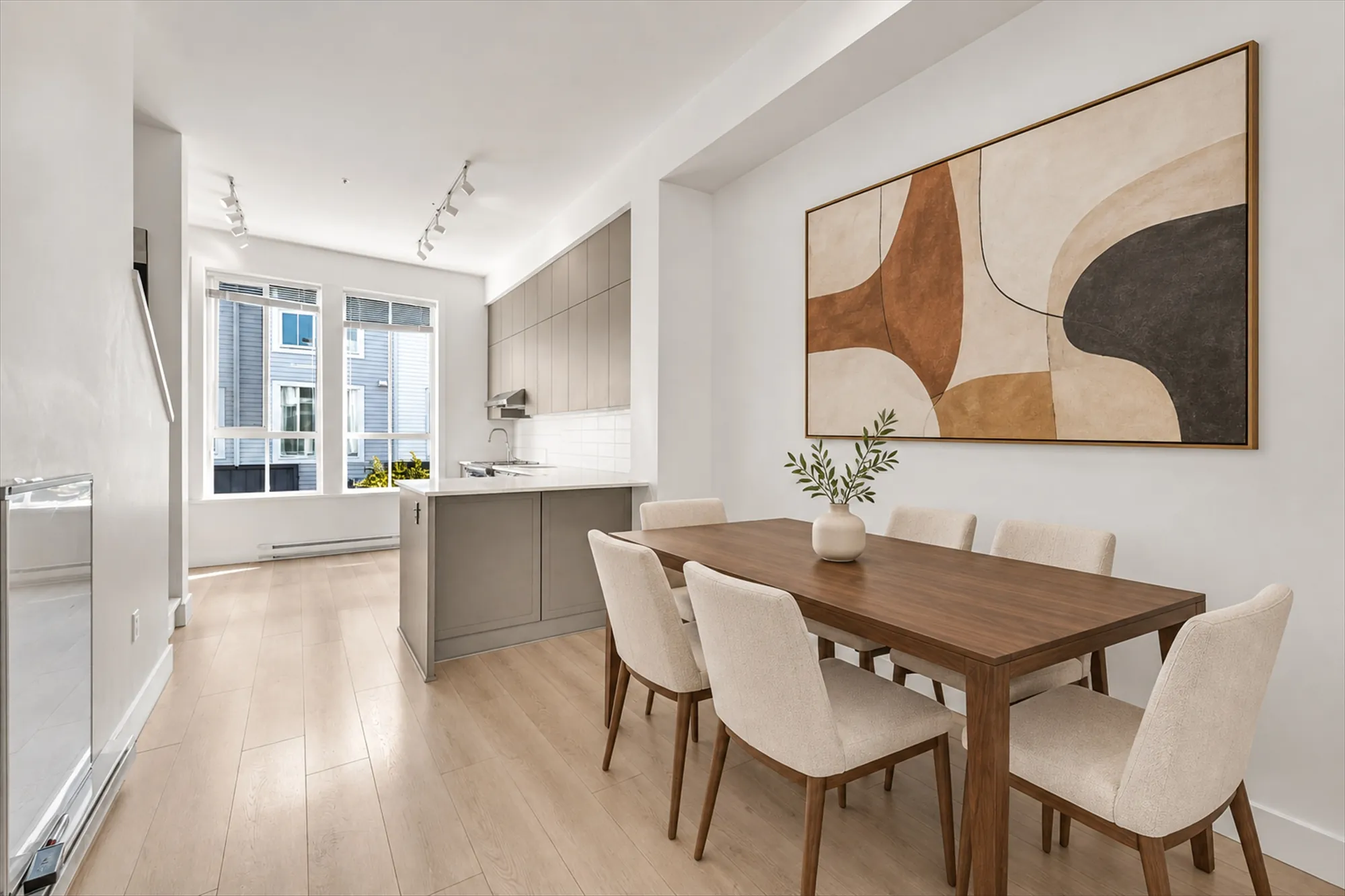 The dining area virtually staged: a long wood table with six cream chairs under an abstract painting, a vase of branches, the kitchen peninsula and the front windows beyond
