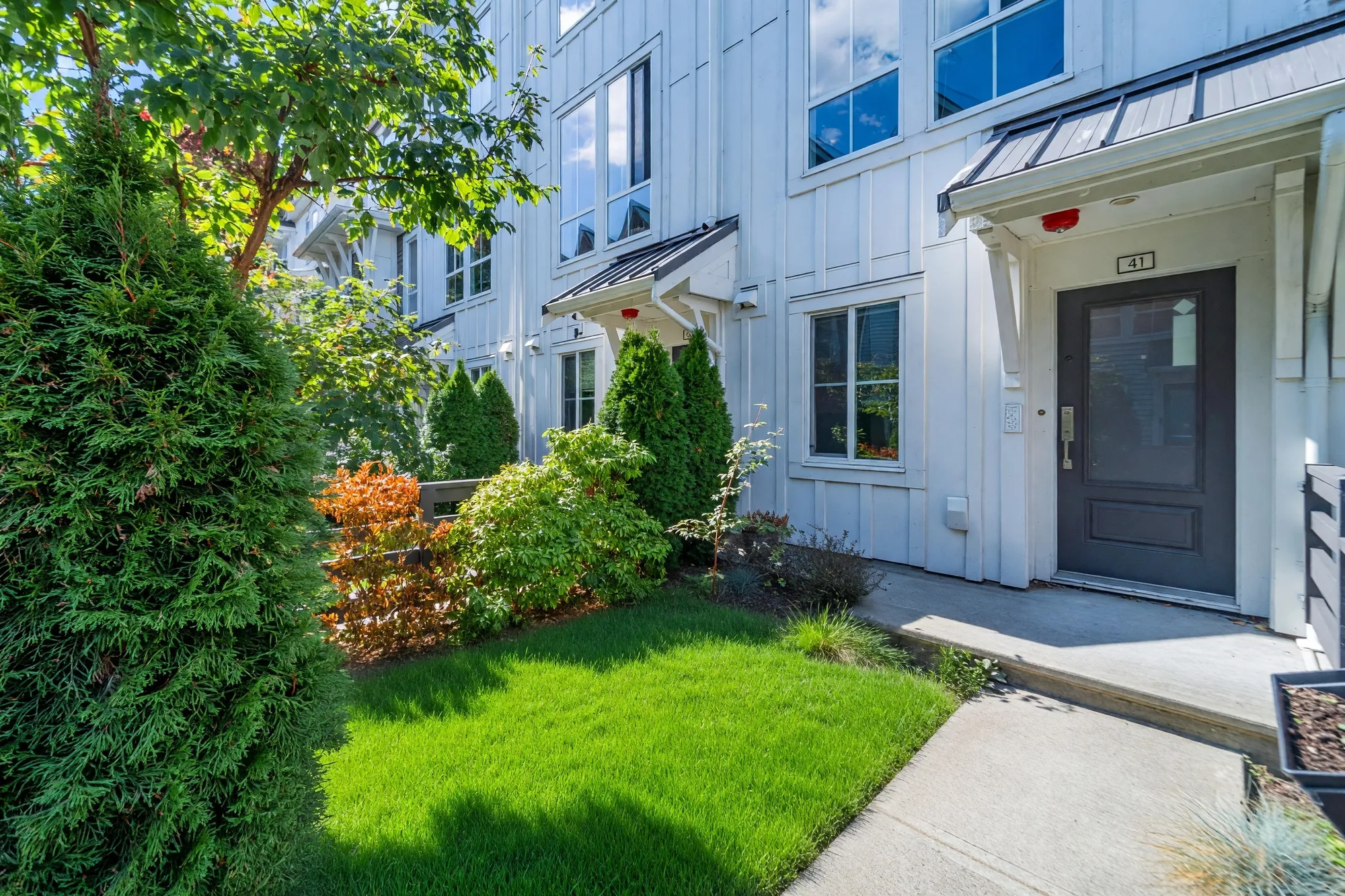 41's front door under its small metal canopy, a patch of lawn shown green, shrubs and a cedar beside the path