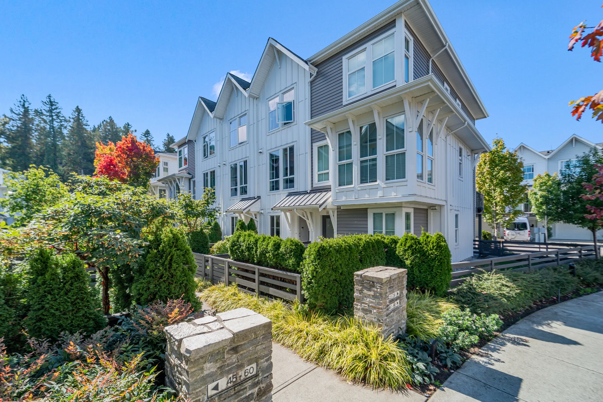The row from the corner of the front walk: white board-and-batten townhouses under gables, a stone pier with a unit-number sign, hedges, grasses and a Japanese maple along the walk