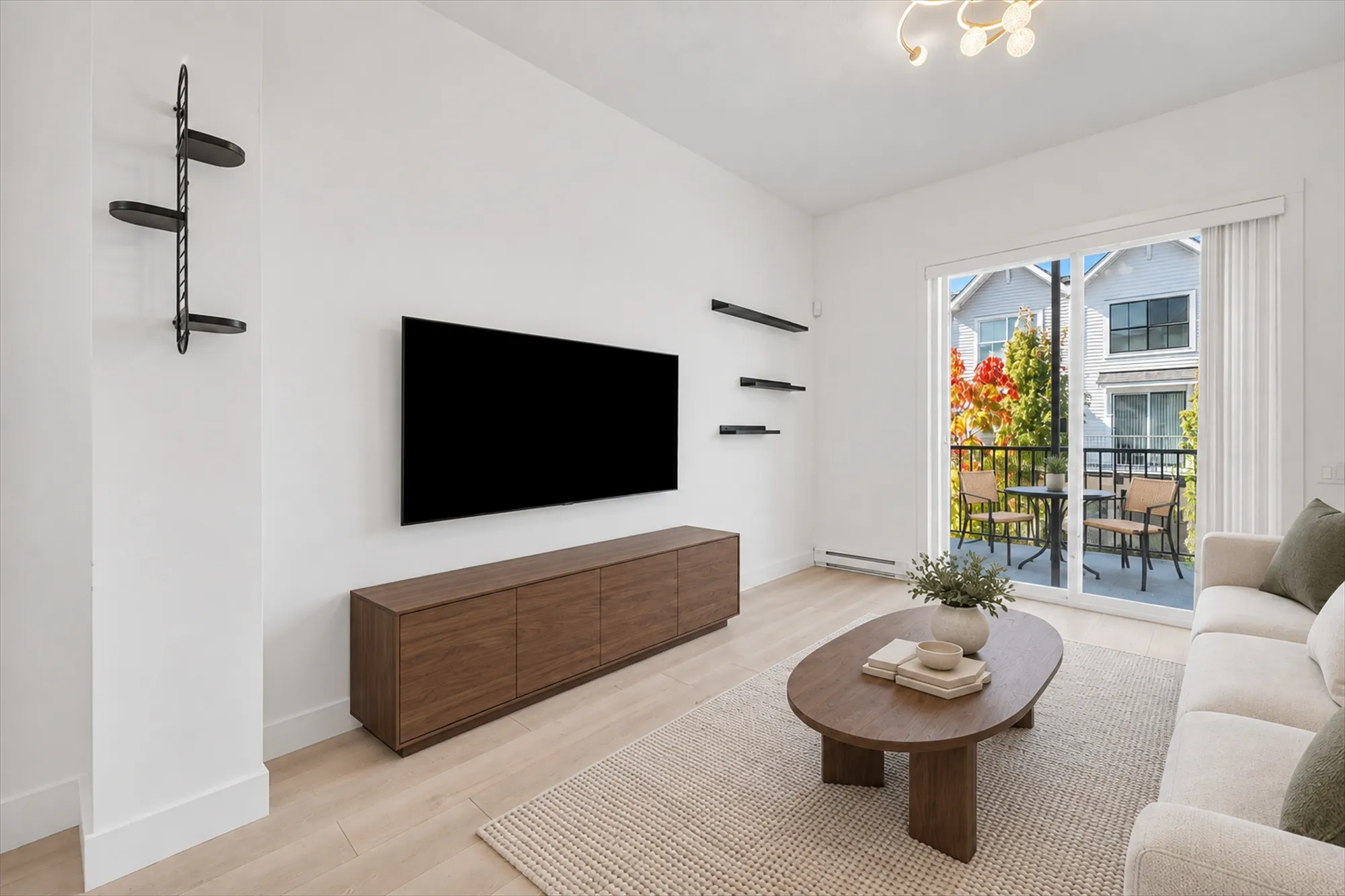 The TV wall virtually staged: a walnut console under the wall-mounted TV, black floating shelves and a wall rack, an oval coffee table on a jute rug, a sofa at the right, a table and chairs on the balcony beyond the sliding door