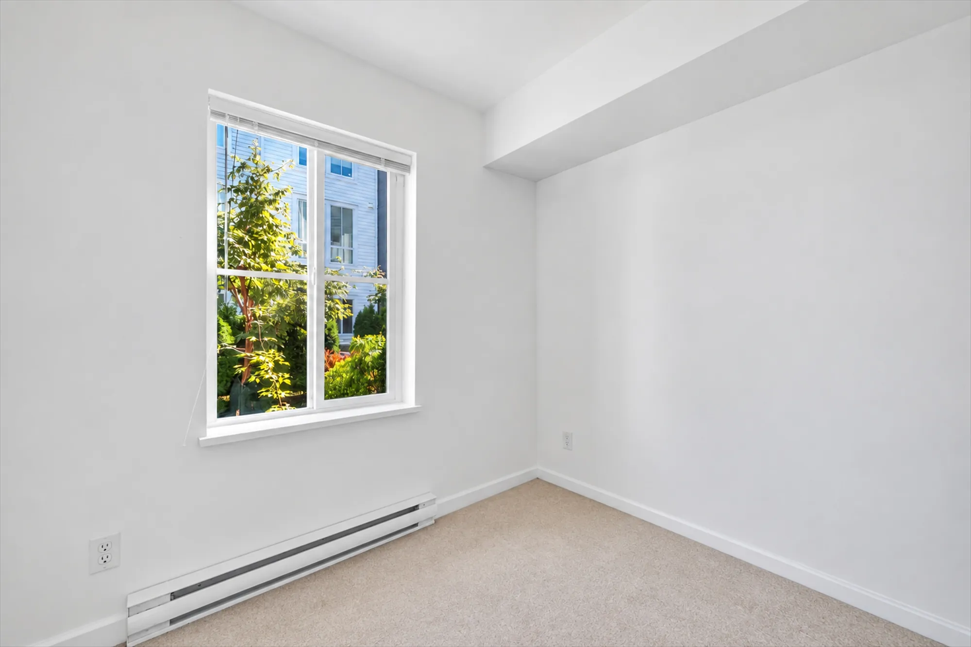 The office shown empty: a window to the front walk, a baseboard heater, carpet, a bulkhead along the ceiling