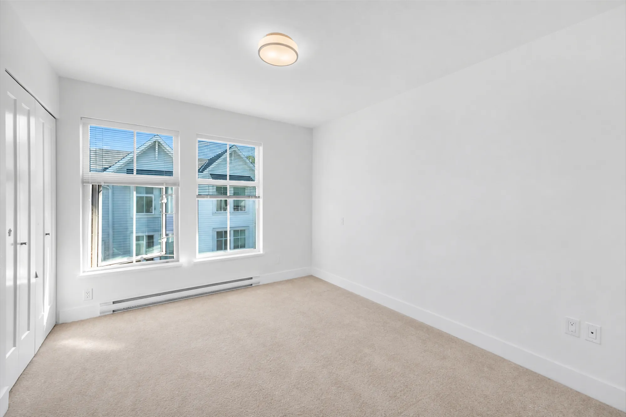 The primary bedroom shown empty: two windows to the front over a baseboard heater, a closet at the left, carpet, a ceiling light