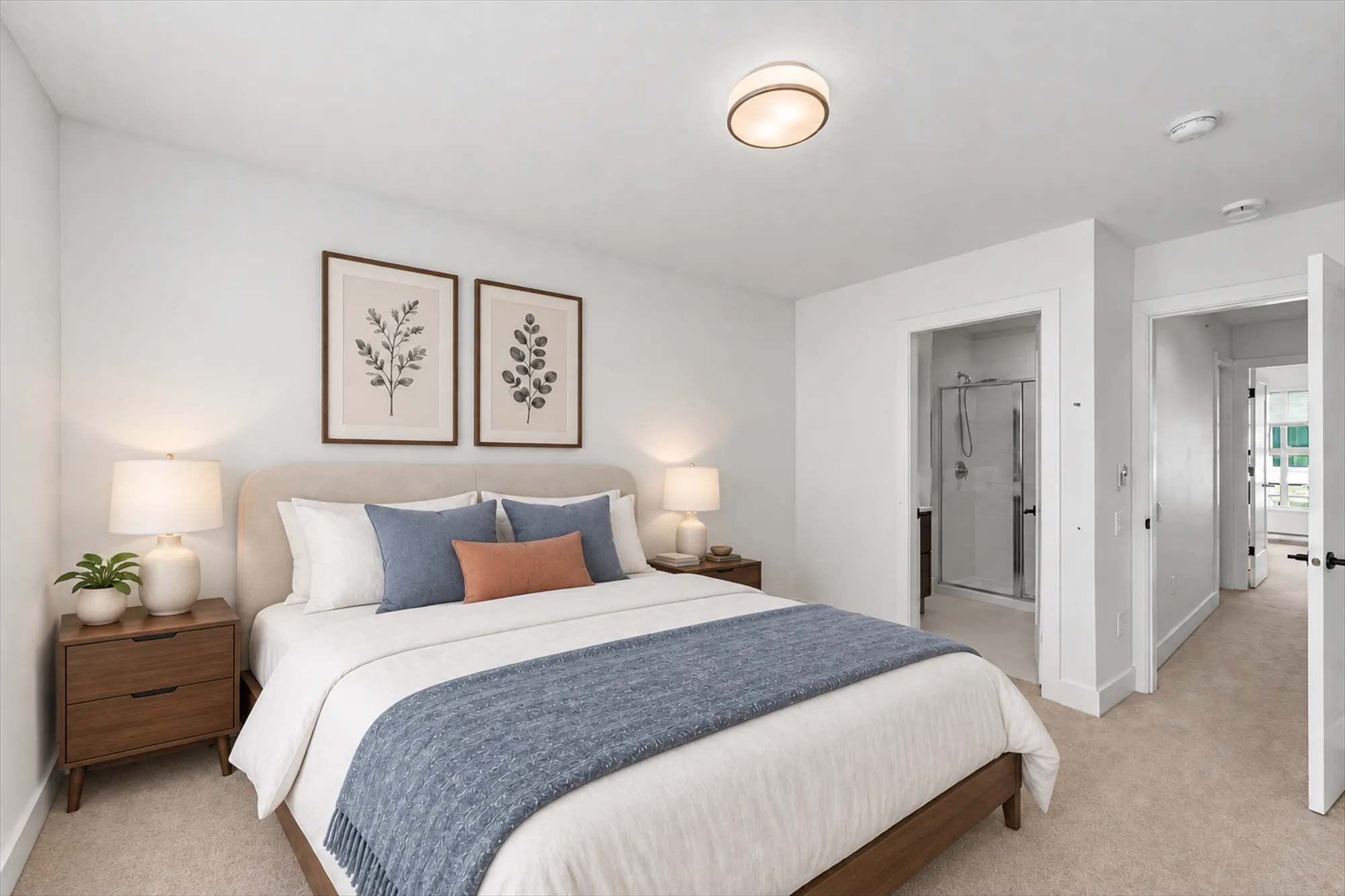 The primary bedroom virtually staged, toward the door to its bathroom with the glass shower beyond and the door to the hall: a bed with white and blue linen, night tables, lamps, two botanical prints