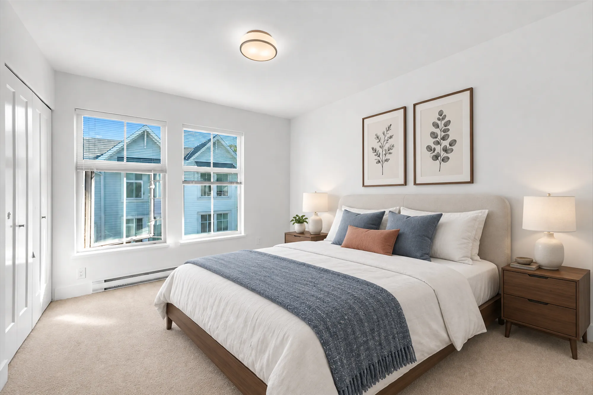 The primary bedroom virtually staged: two windows to the front over a baseboard heater, a bed with white and blue linen between two night tables with lamps, two botanical prints, a closet at the left